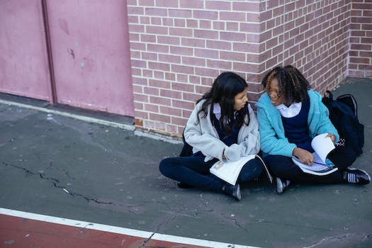 From above of diverse clever cute classmates studying information from textbooks and sitting on ground near corner of brick building