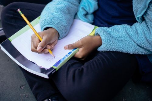Free Crop anonymous ethnic child writing on sheet of paper with pencil while sitting on ground Stock Photo