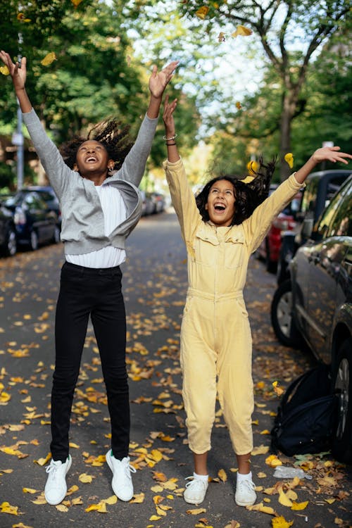 Full body of cheerful cute multiethnic girls with raised hands looking up and screaming on street with autumn leaves