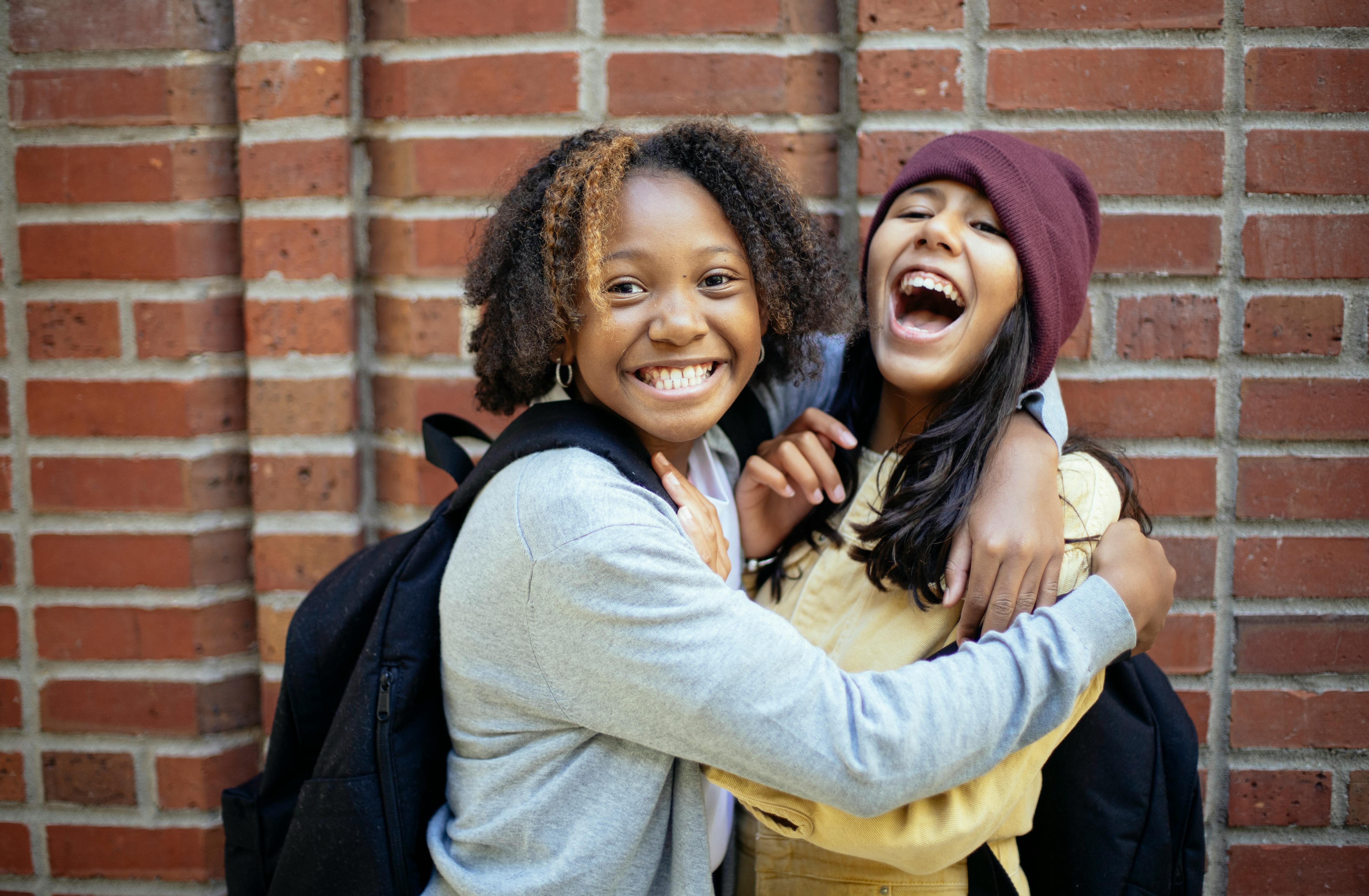 cheerful diverse schoolgirls embracing near brick wall