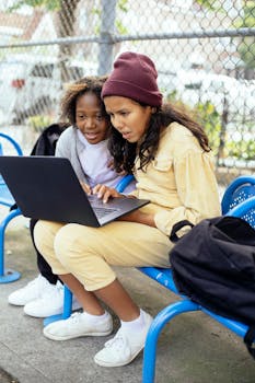 Two diverse schoolgirls using a laptop on a bench outdoors, enjoying technology together.