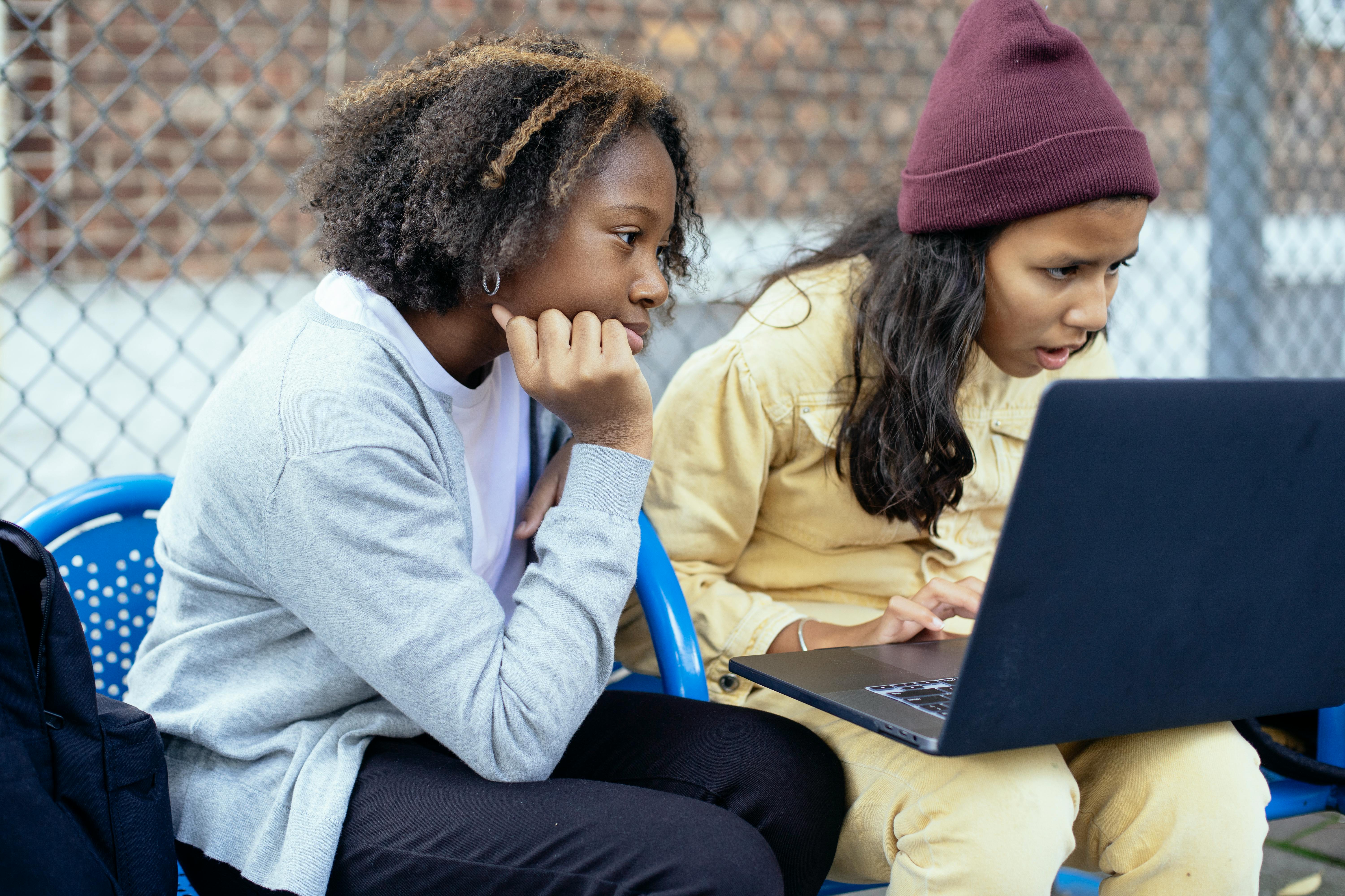 attentive diverse schoolgirls watching laptop on street bench