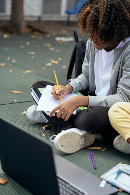 Focused African American schoolchild taking notes on paper while doing homework with crossed legs near netbook and unrecognizable friend on pavement