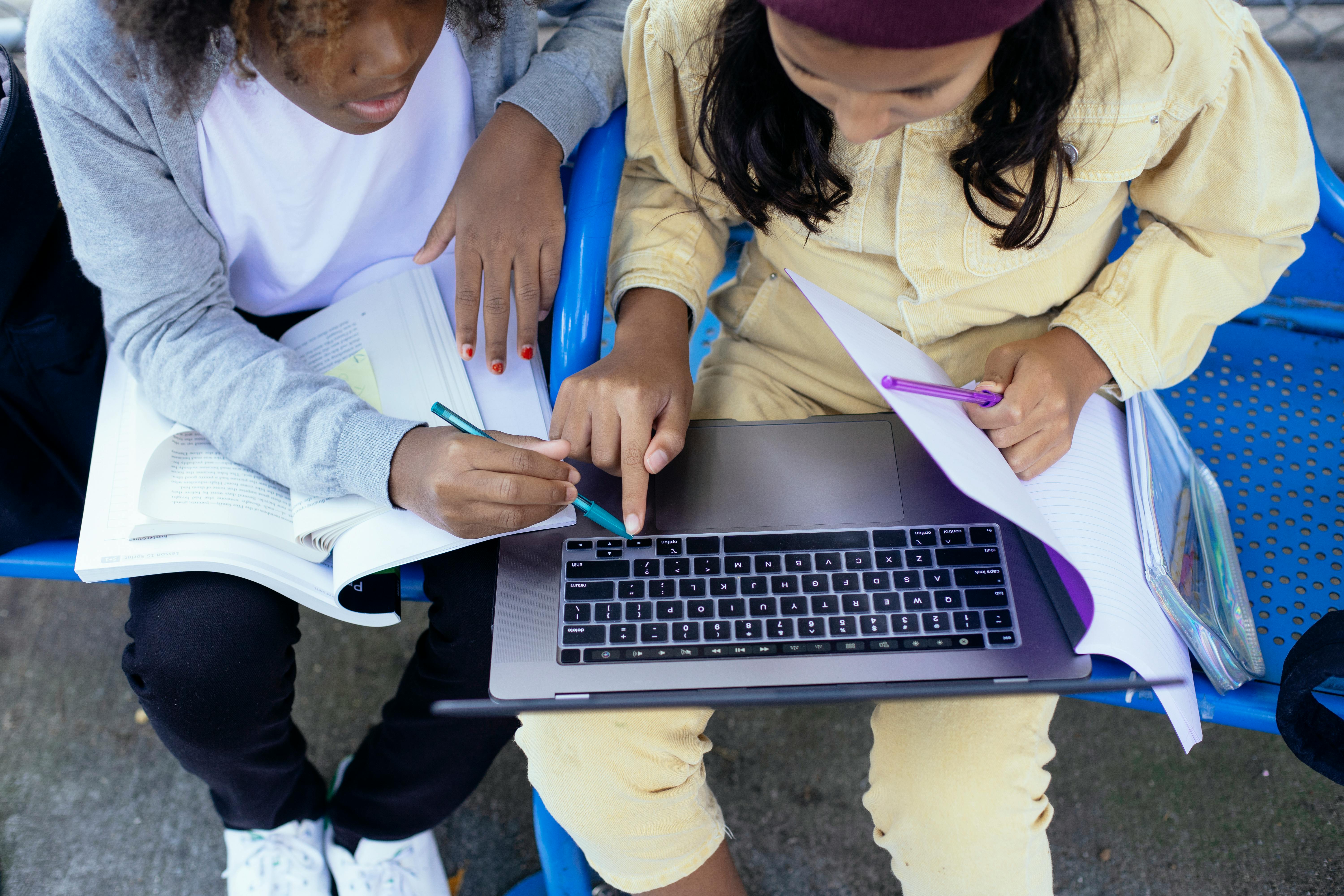 crop multiethnic schoolgirls with laptop and workbooks doing homework outdoors