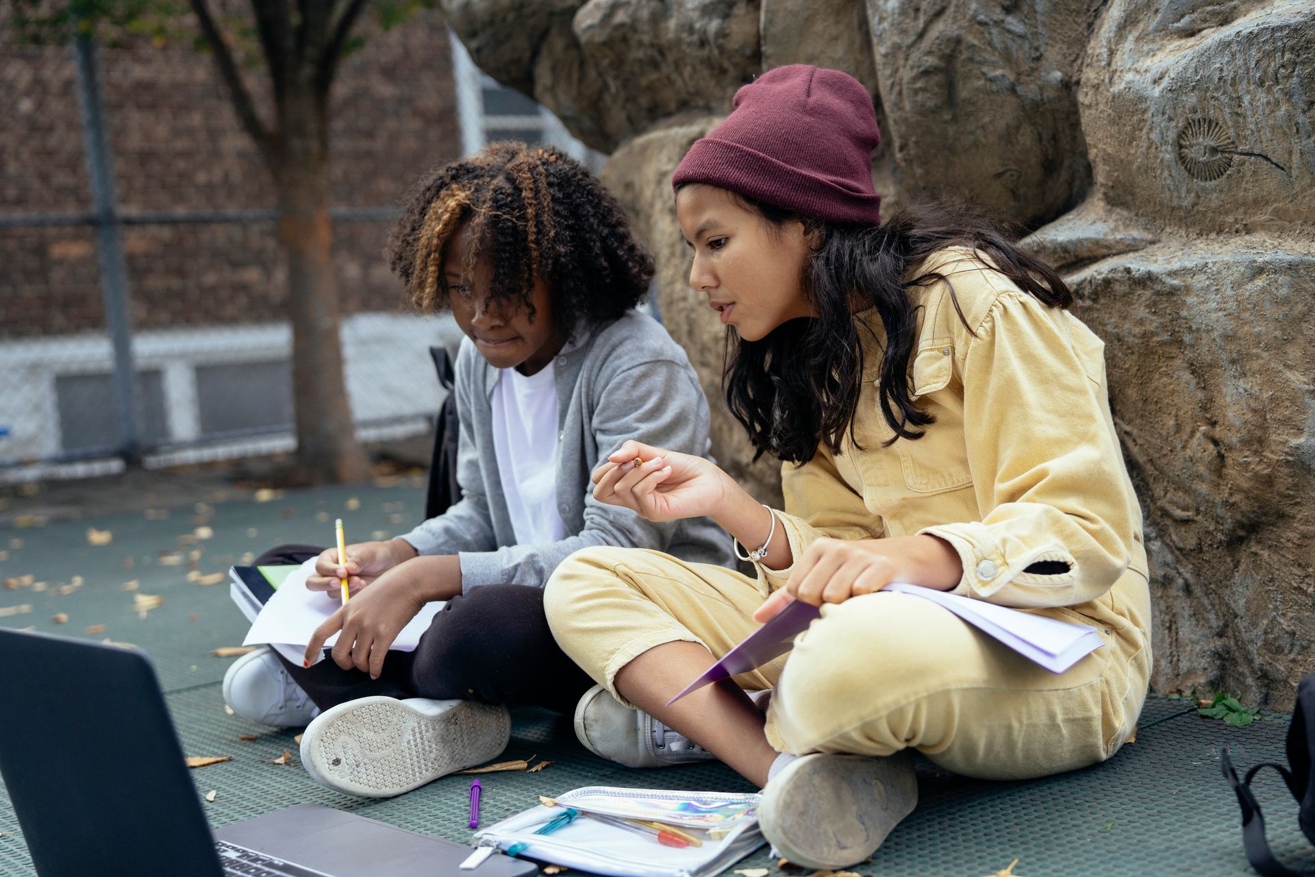 Multiracial schoolchildren with workbooks interacting while watching netbook and sitting with crossed legs on street