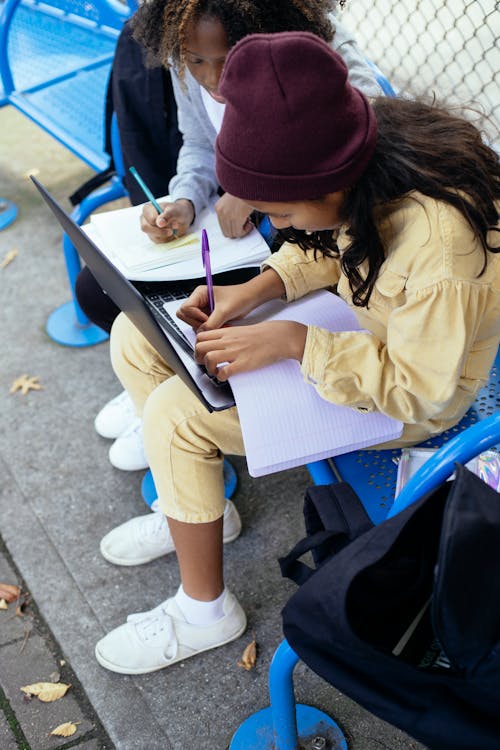 High angle of crop anonymous multiracial schoolkids with netbook writing in workbook while doing homework on urban bench