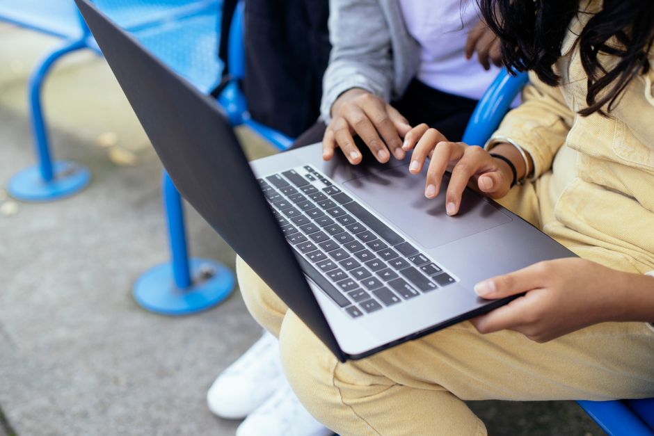 Crop anonymous diverse schoolchildren browsing internet on netbook while resting on urban bench in daylight