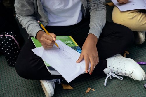 Free From above of crop ethnic schoolkid drawing with pencil on paper sheet while sitting with crossed legs on pavement near anonymous friend Stock Photo