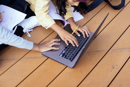 Crop ethnic schoolchildren typing on laptop while studying on street