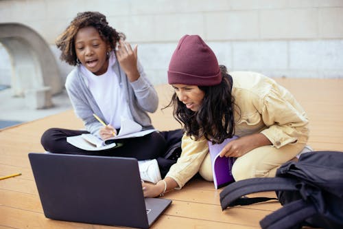 Free Attentive diverse schoolchildren watching portable computer while doing homework and talking on wooden platform in daylight Stock Photo