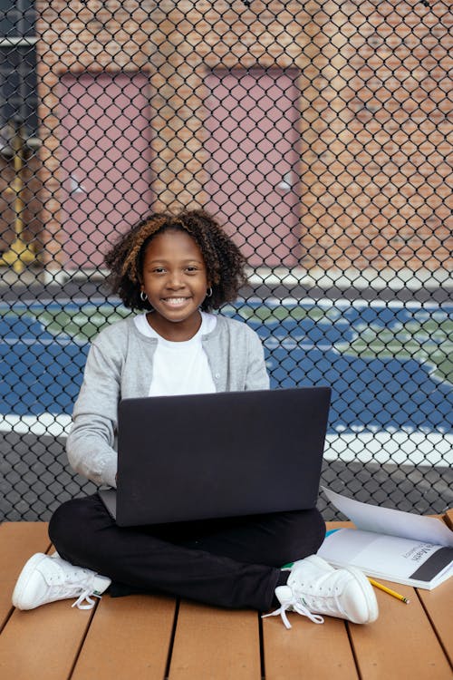 Content black schoolchild with laptop studying near grid fence