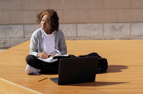 Free Contemplative African American schoolkid with copybook and netbook sitting with crossed legs on wooden platform while looking away in sunlight Stock Photo