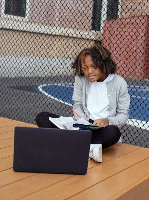 Attentive black schoolkid with exercise book watching netbook while doing homework with crossed legs near grid fence