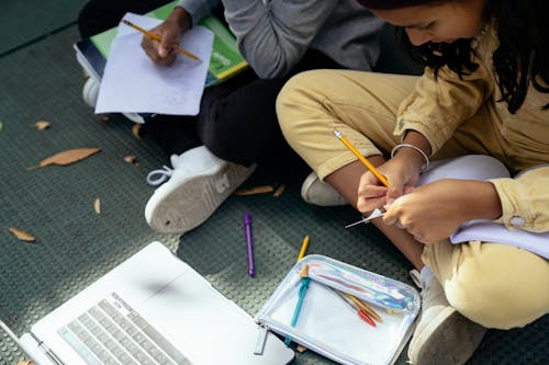 Crop Hispanic schoolgirl writing in copybook near unrecognizable friend outdoors