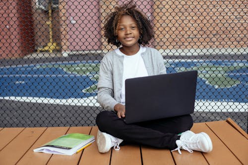 Smiling African American schoolchild with Afro hairstyle sitting with netbook on crossed legs while studying and looking at camera near grid fence