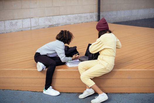 Side view of anonymous multiracial schoolkids with workbook and netbook doing homework on wooden platform