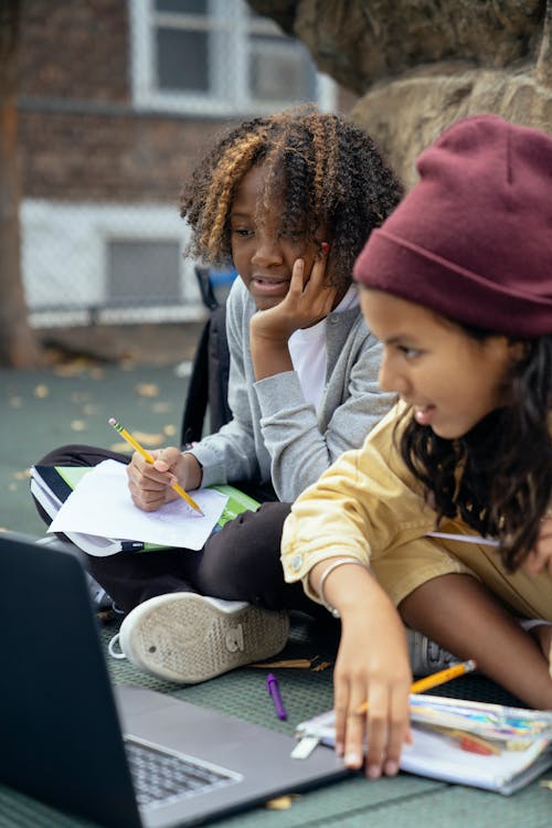 Crop glad multiethnic schoolkids sitting with crossed legs while watching netbook and doing homework on urban pavement