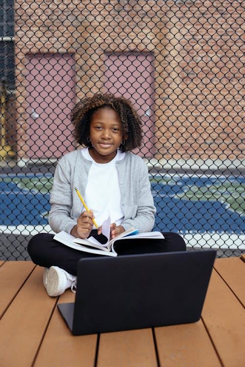 Happy African American schoolgirl sitting on wooden surface with laptop and notepad while preparing for exam and looking at camera