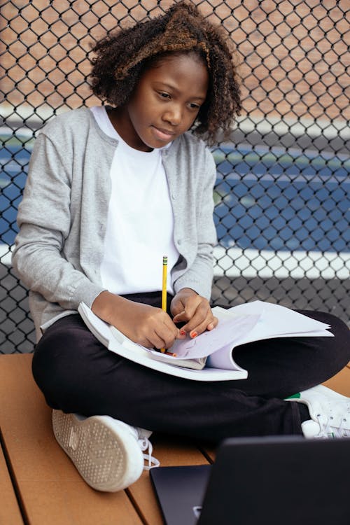 Free African American kid with curly hair sitting with crossed legs and writing notes while looking at screen of laptop Stock Photo