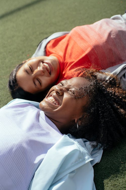 High angle of cheerful multiracial girlfriends lying on green lawn and toothy smiling while having fun together