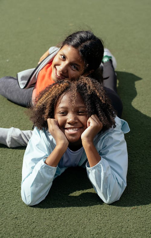 Cheerful diverse girls classmates resting on lawn while spending time together under sunlight