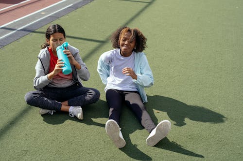 Full body of diverse girlfriends in sportswear sitting on lawn while drinking water after workout together