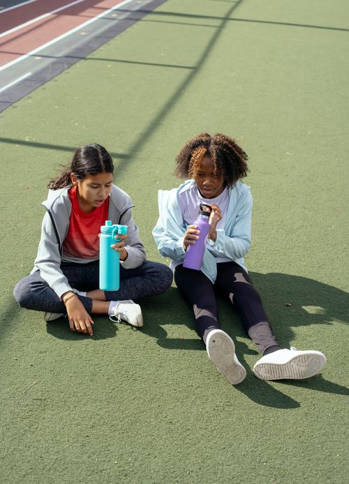 Free Full length of multiracial schoolgirls in activewear resting on stadium with bottles of water during break from workout Stock Photo
