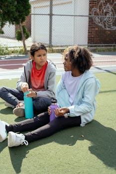 Diverse schoolgirls sitting on stadium with plastic bottles of water while relaxing after training