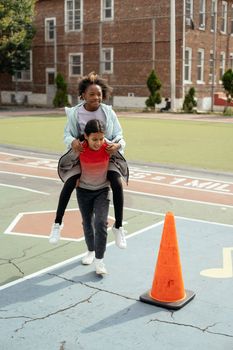 Happy Hispanic kid wearing sportive outfit piggybacking African American classmate while running along backyard school playground on sunny day