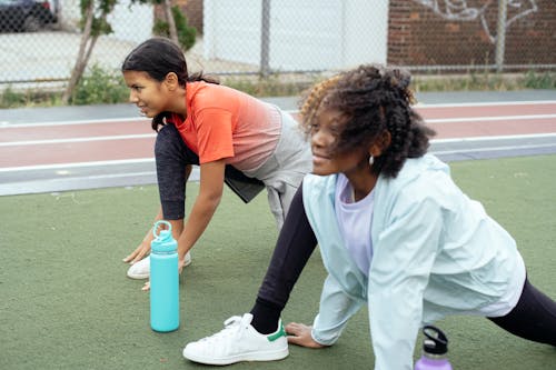 Side view of smiling multiracial sportive kids wearing activewear warming up legs before running training on outdoors track in daytime