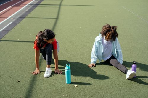 Free From above African American girl doing split and communicating with friend stretching legs on green surface with sport water bottles near track on sunny day Stock Photo