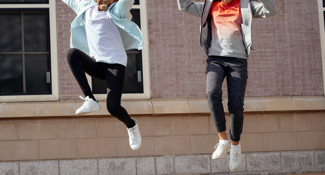 Free Crop smiling black girl and unrecognizable classmate wearing sportive clothes jumping high together while spending free time after lessons near school Stock Photo