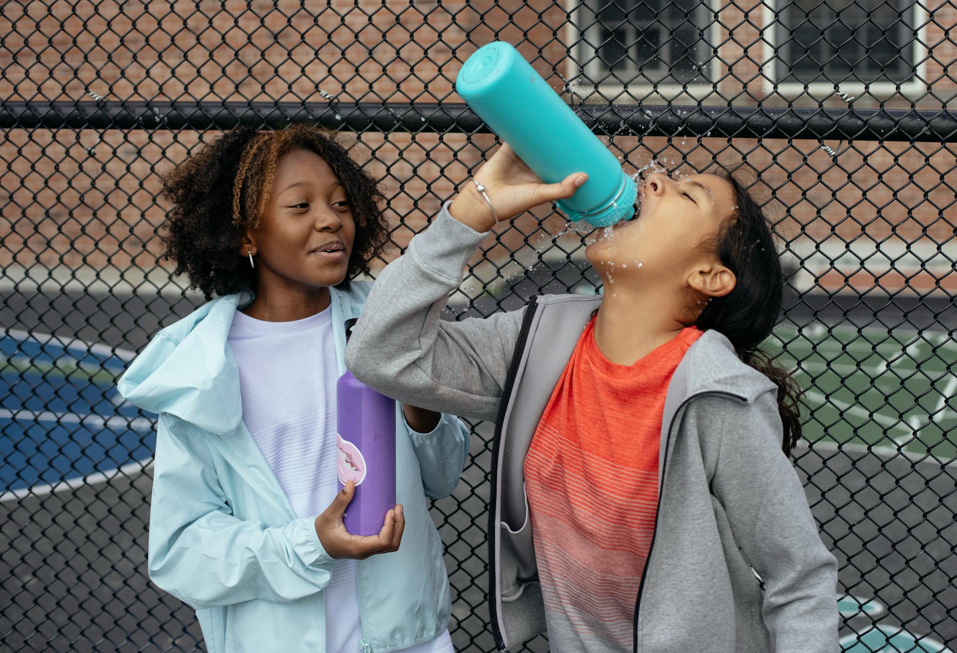 Thirsty multiracial girls in sportswear drinking water from plastic bottle during break in training on sports ground