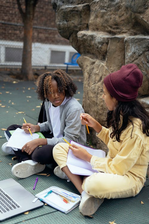 Free Concentrated diverse girls in casual outfits sitting with copybooks and laptop on ground while doing homework together Stock Photo
