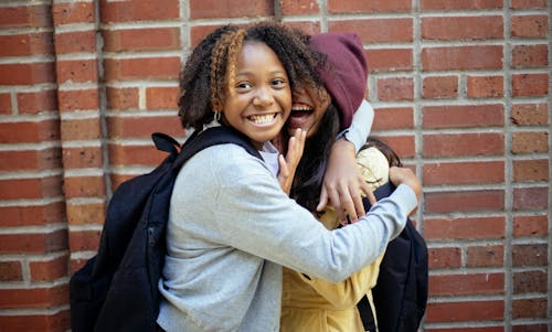 Cheerful African American girl in casual clothes hugging friend while having fun near brick wall