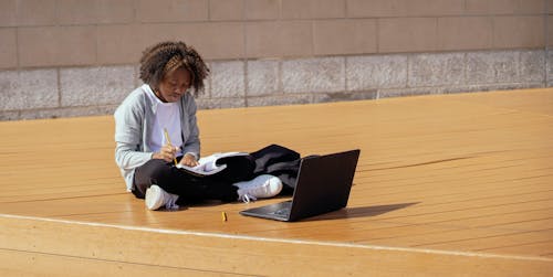 Full body of focused African American girl sitting with laptop and copybook and doing homework