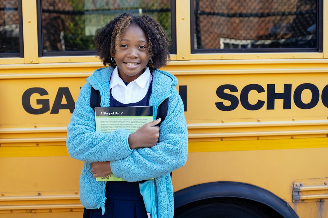 Free Cheerful African American girl in school uniform and warm jacket standing with textbook near yellow school bus and looking at camera Stock Photo
