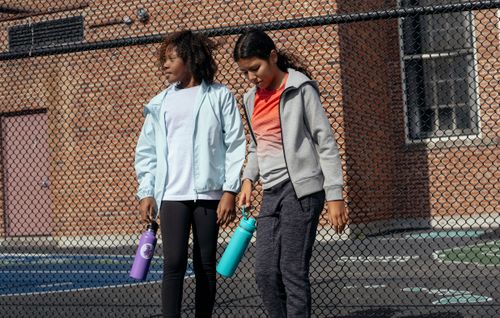 Free Tired multiracial girls in sportswear with plastic water bottles standing near metal net fence while having break in training Stock Photo