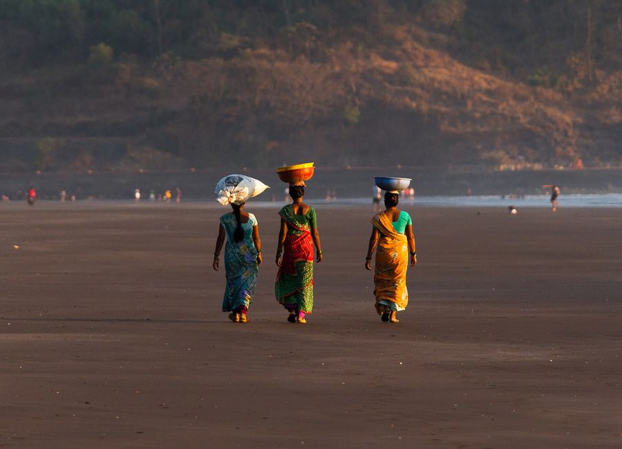 Three women in colorful sarees walk along Harihareshwar Beach in India, balancing items on their heads.