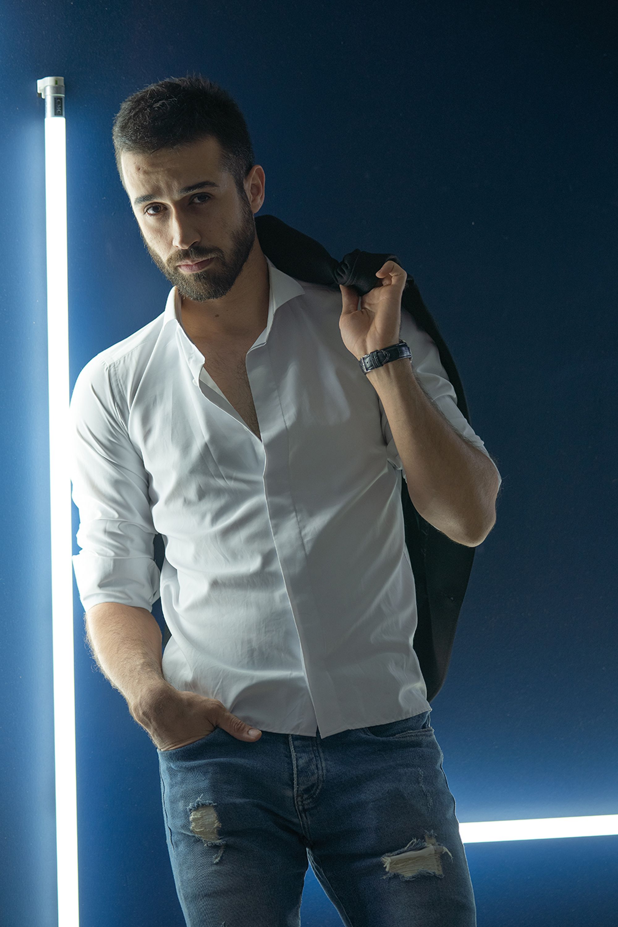 Portrait of a young man in Tehran wearing a white shirt and jeans, backlit by fluorescent lights.