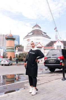 A stylish woman in a hijab stands confidently in front of a historic Bandung building.