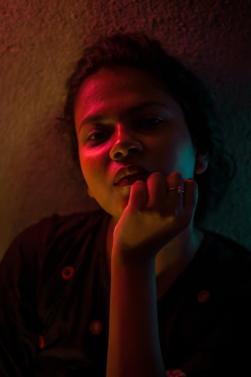 Pensive young Indian female with dark hair lean on on wall with hand at chin and looking at camera in dark room with neon lights