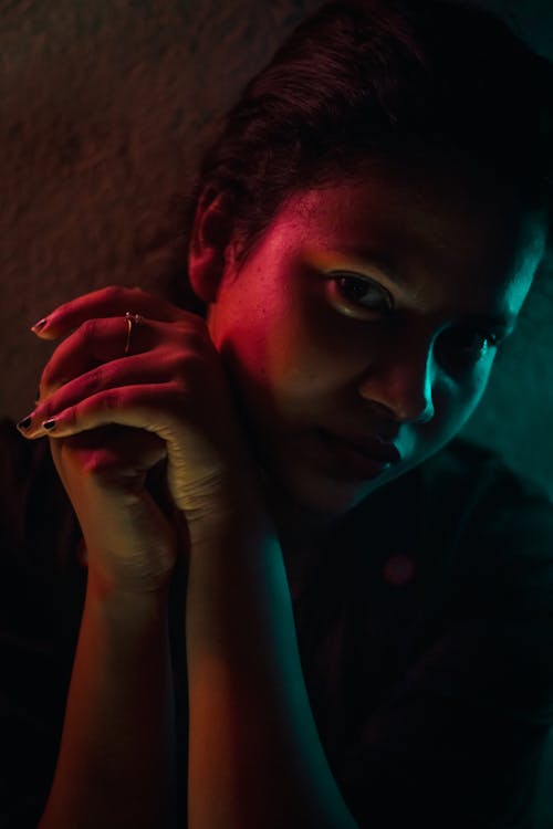 Crop young dreamy Indian lady sitting leaning on hands and looking at camera in dark room with colorful neon lights