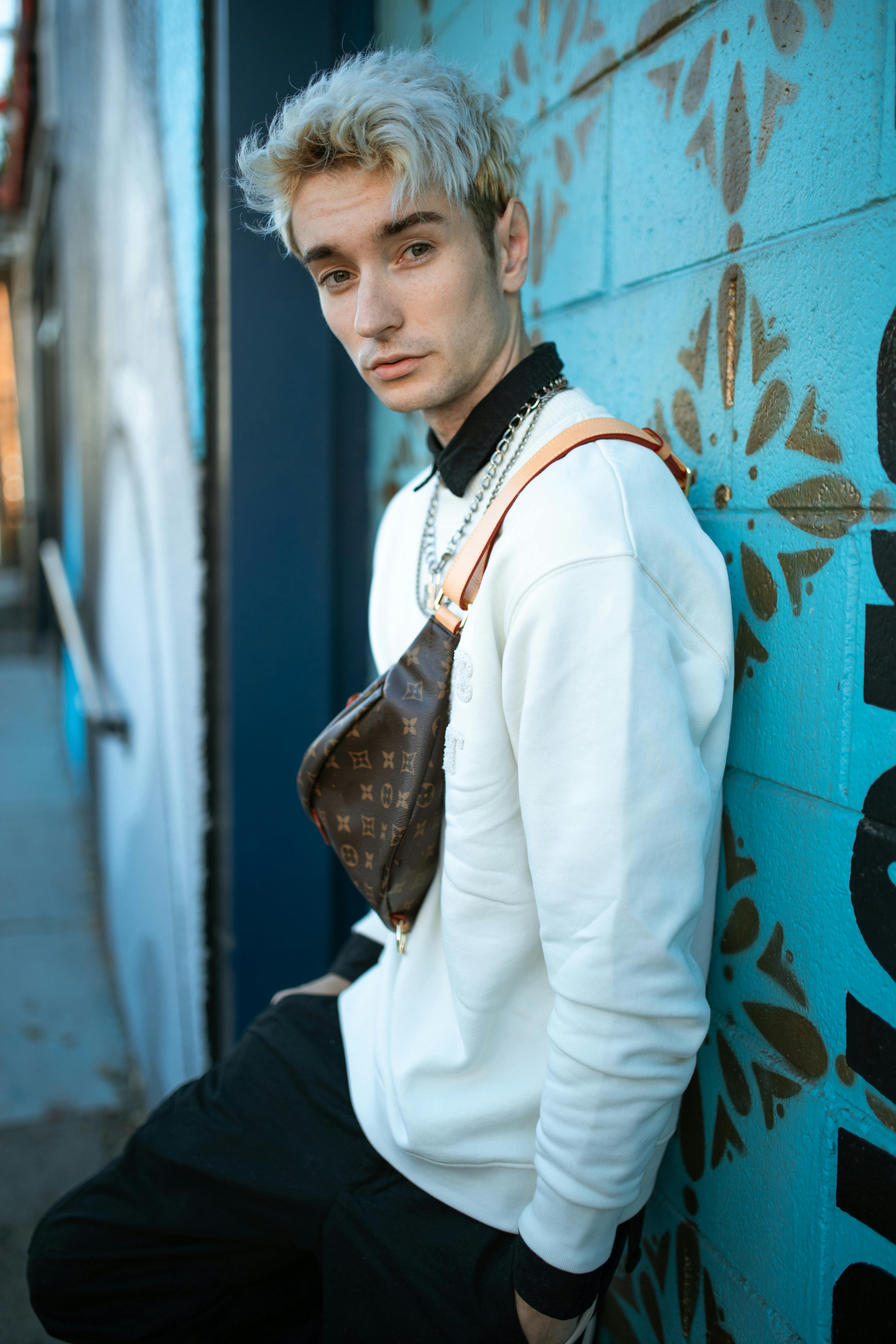 Portrait of a young man with trendy dyed hair and fashionable sling bag against an urban graffiti backdrop.