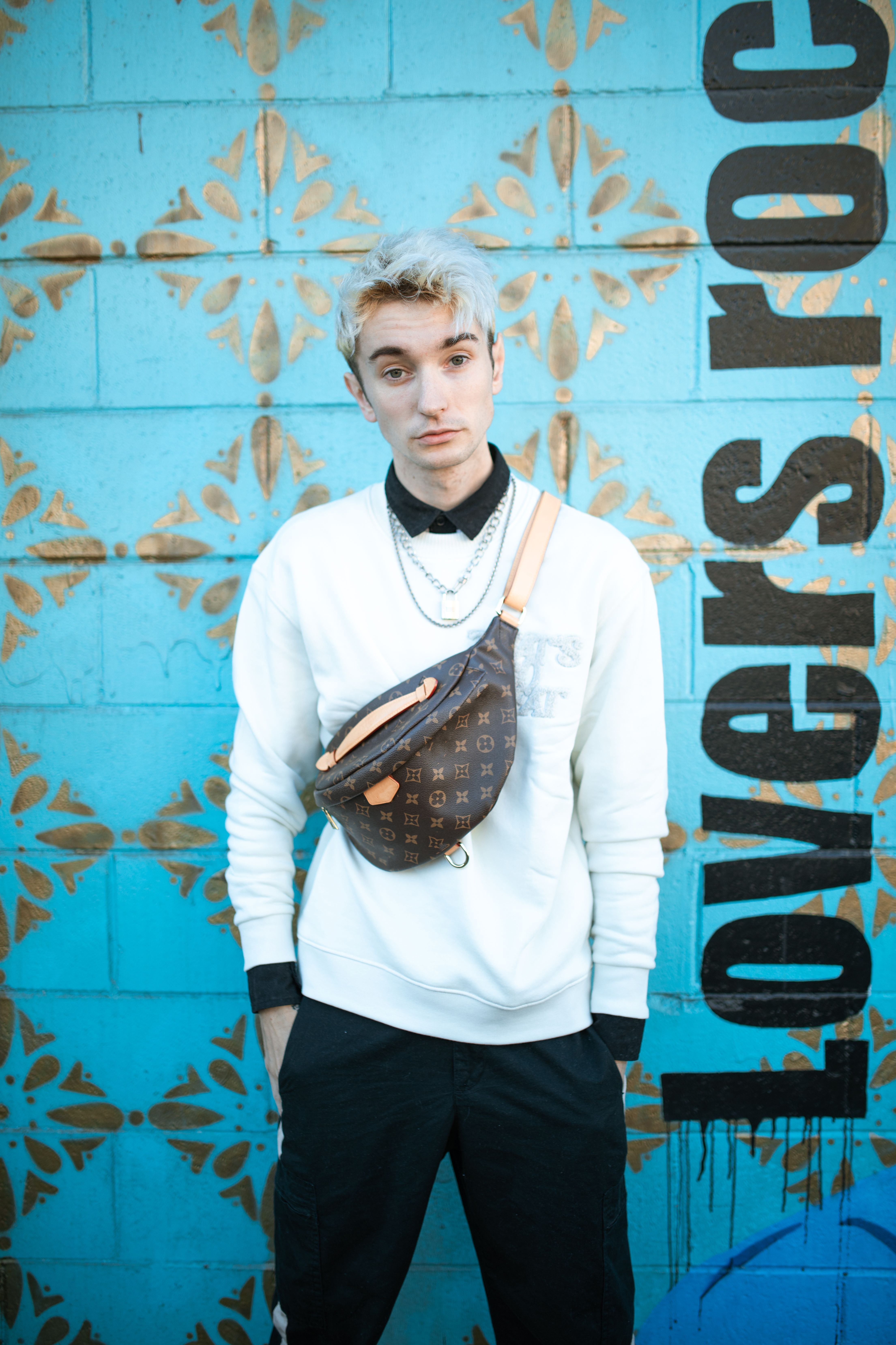 Fashion-forward young adult poses against blue graffiti wall, showcasing street style with a sling bag.
