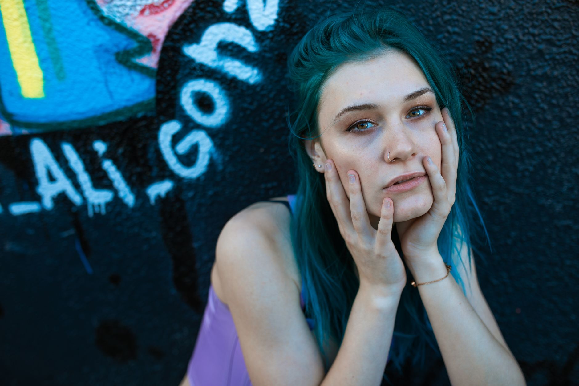 Artistic portrait of a woman with piercings and blue hair posed against graffiti.