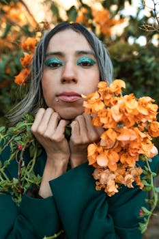 A person poses amidst orange flowers with bold makeup, showcasing unique style and self-expression.