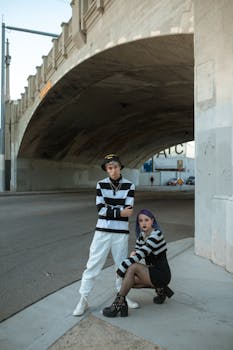 Trendy Gen Z duo in striped outfits posing under a city bridge.