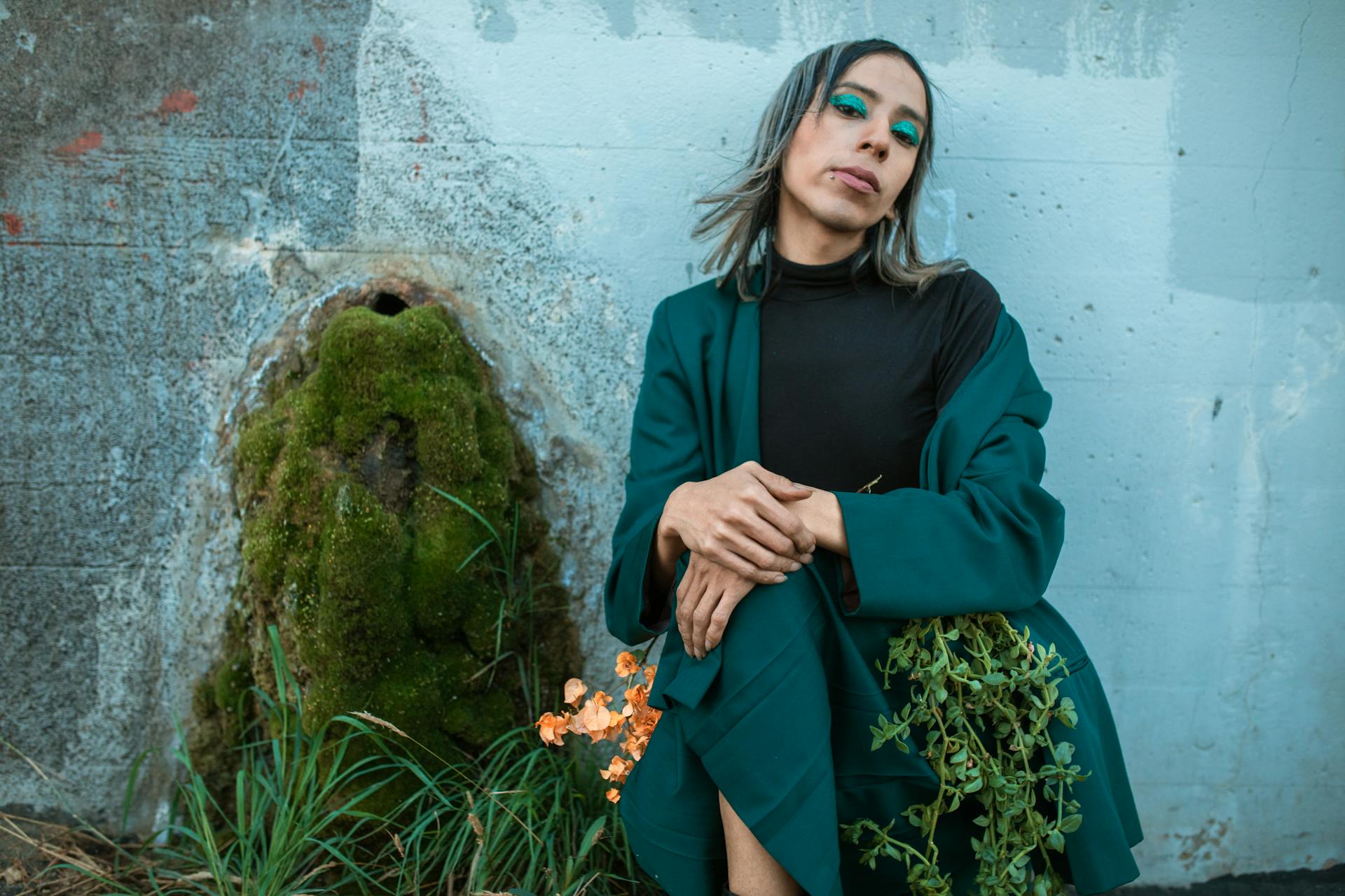 Stylish androgynous individual posing against a wall with greenery.