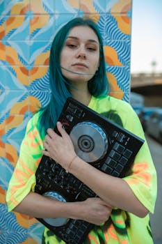 Young DJ girl with blue hair, holding a controller against vibrant tiles. Trendy street style.